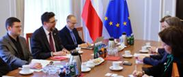 Po dwóch stronach czworokątnego stołu siedzi kilka rozmawiających osób, za stołem pod ścianą flagi Polski i UE.