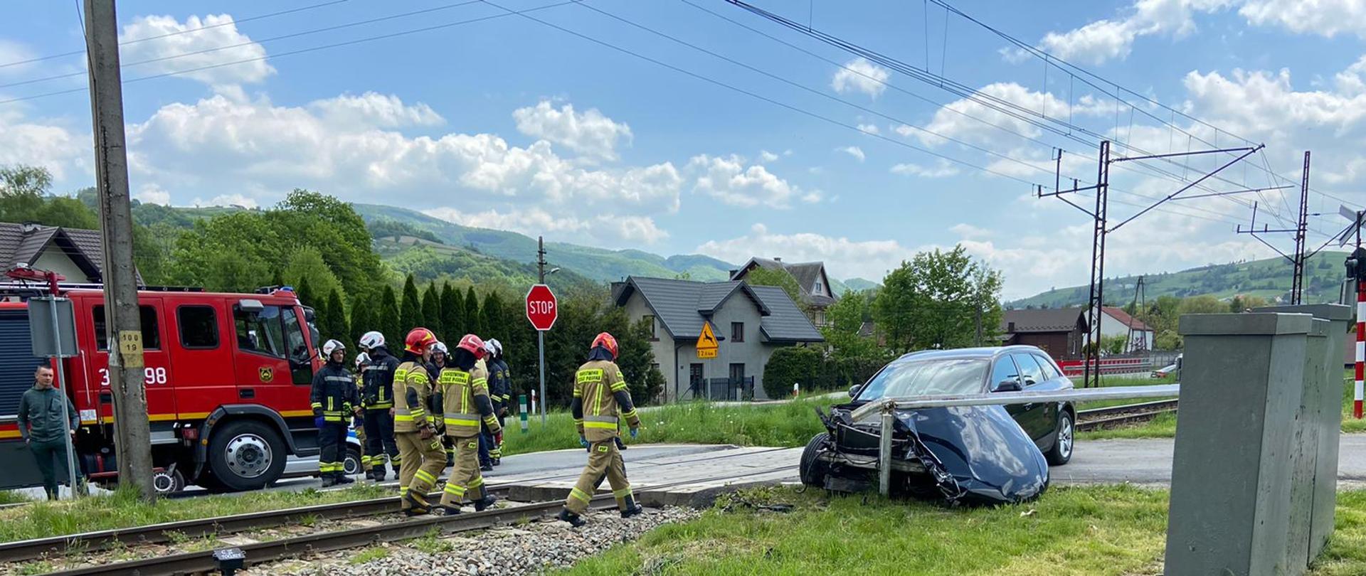 Uszkodzony samochód osobowy na przejeździe kolejowym. Obok strażacy.