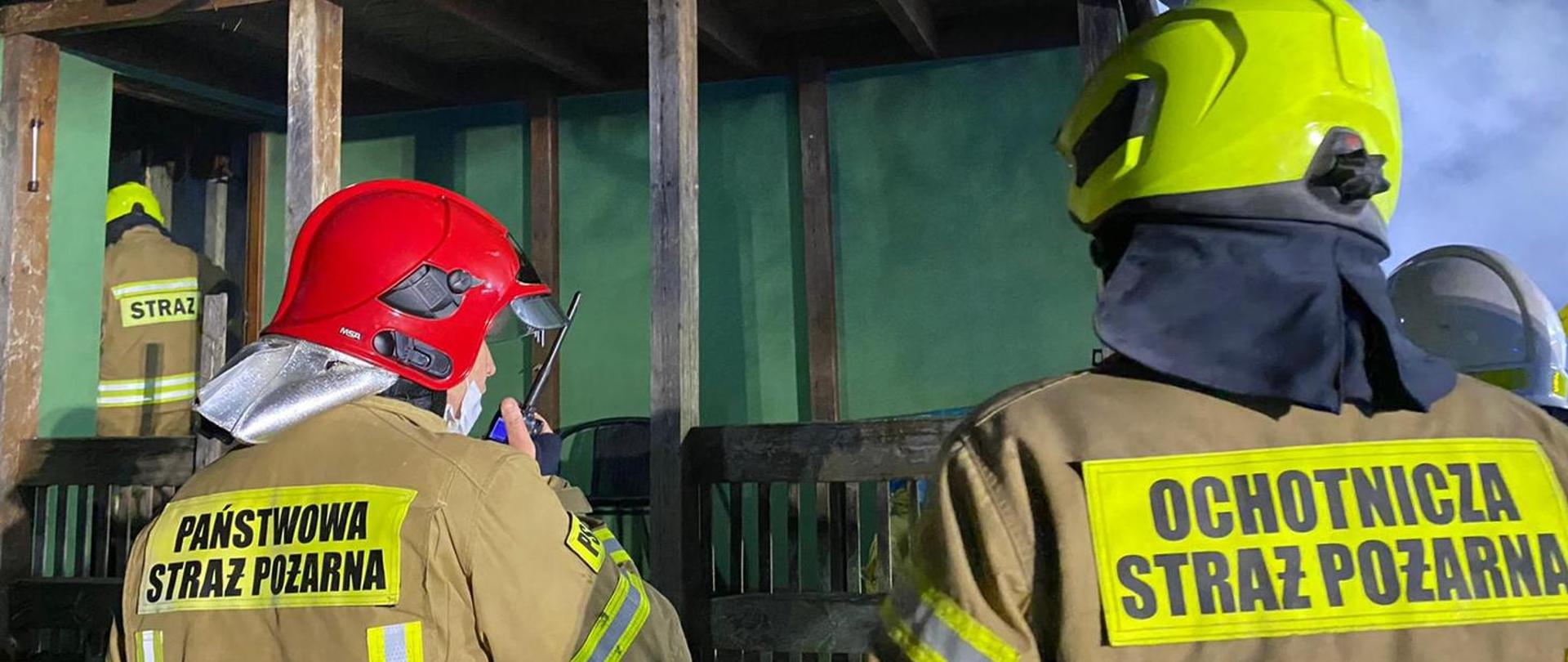 Zdjęcie przedstawia dwóch mężczyzn- strażaków z Państwowej Straży Pożarnej i Ochotniczej Straży Pożarnej w umundurowaniu typu nomex. W tle budynek z zieloną elewacją z drewnianymi elementami.