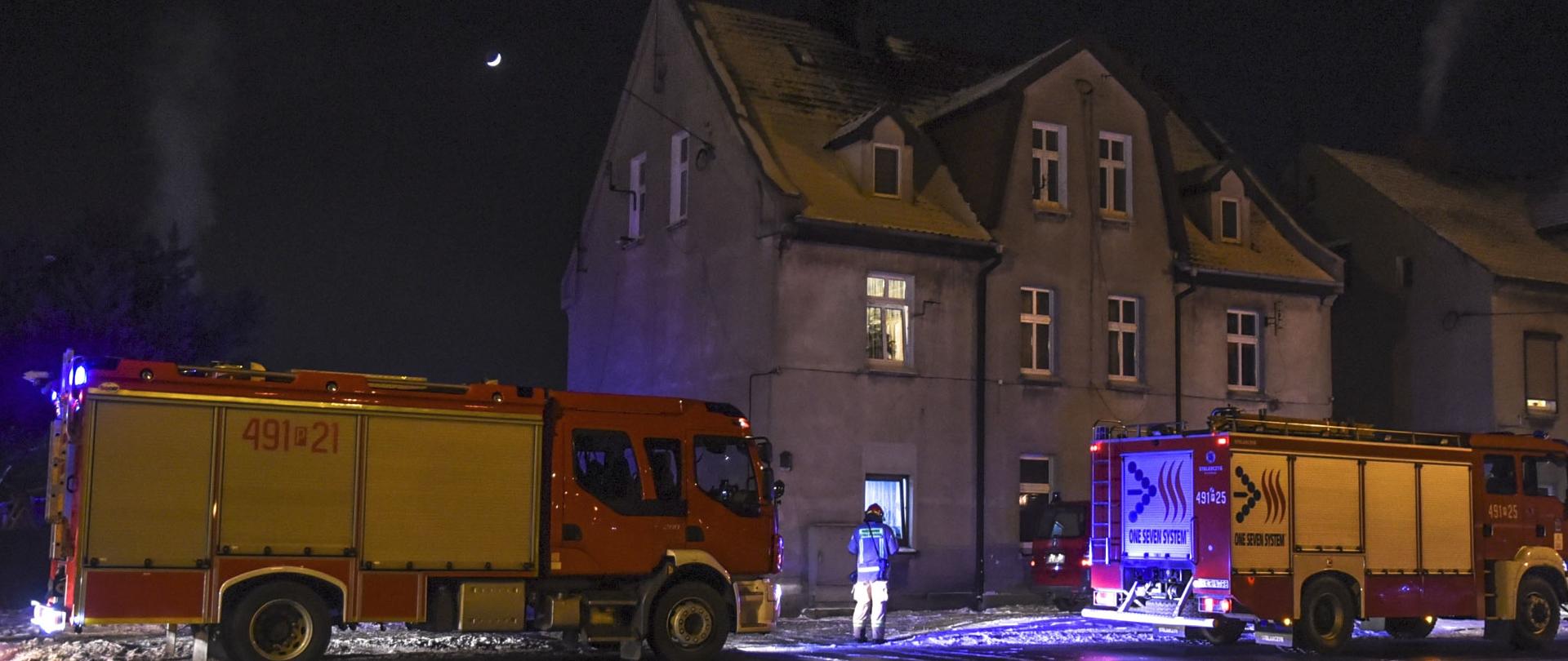 Na tle budynku wielorodzinnego stoją samochody pożarnicze. Strażacy podejmują działania związane z weryfikacją zagrożenia pod kątem występowania tlenku węgla. Jest noc.