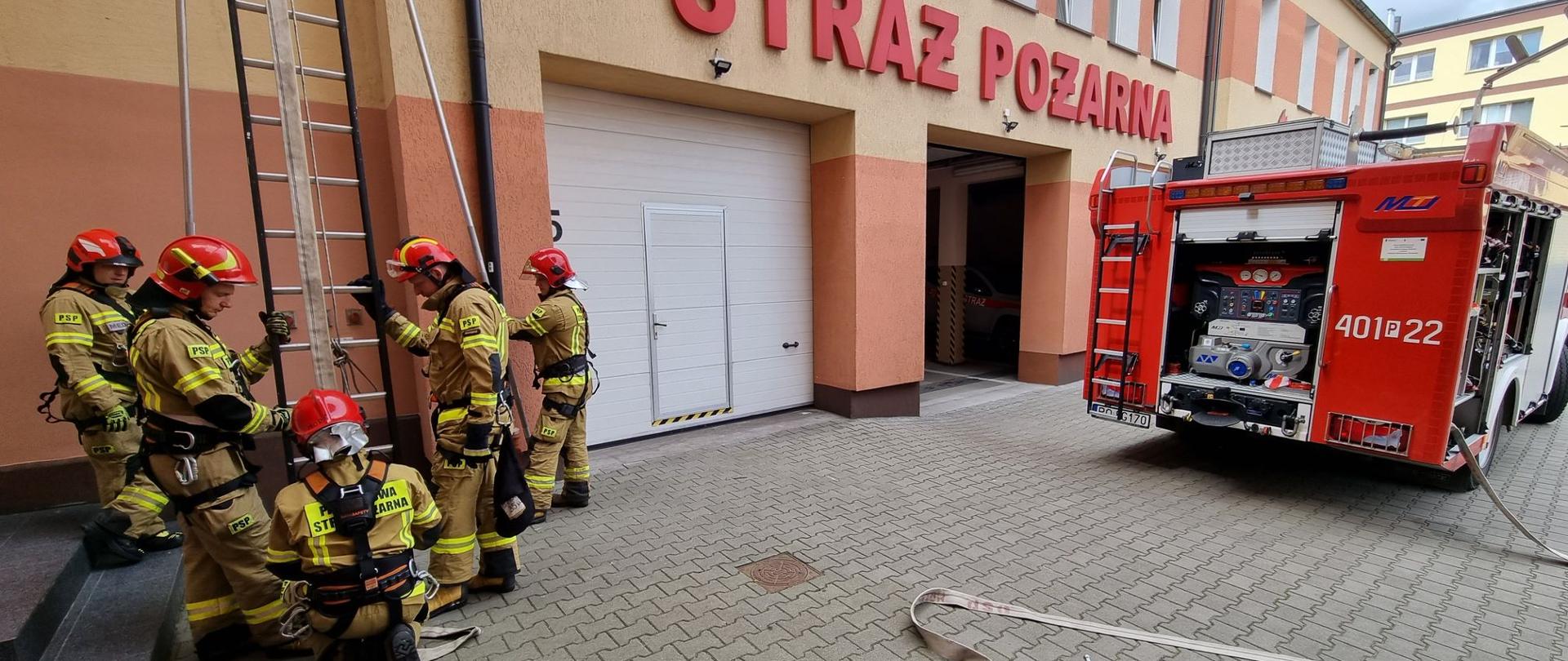 Zdjęcie przedstawia strażaków ćwiczących w ramach doskonalenia zawodowego.
W tyle plac komendy i budynek jednostki.
