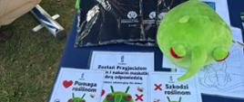 Zielona, pluszowa maskotka leży na stoliku pośród kolorowych kartek dotyczących konkursu dla dzieci "Zostań przyjacielem roślin - nakarm maskotkę dobrą odpowiedzią".
