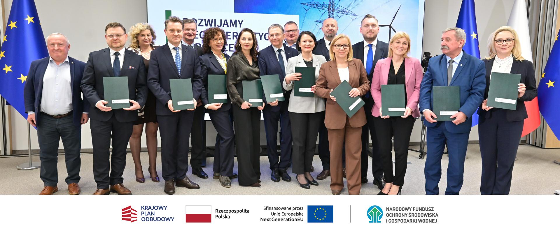 Na zdjęciu: Paulina Hennig-Kloska, minister Klimatu i Środowiska, Konrad Wojnarowski, podsekretarz stanu w Ministerstwie Energii, Dorota Zawadzka-Stępniak, prezes zarządu NFOŚiGW, Józef Matysiak, zastępca prezesa zarządu NFOŚiGW, Paweł Augustyn, zastępca prezesa zarządu NFOŚiGW, Robert Gajda, zastępca prezesa zarządu NFOŚiGW, Dariusz Lubera, prezes zarządu PGE S.A., Jacek Drozd, prezes zarządu PGE Dystrybucja S.A., Marcin Laskowski, wiceprezes zarządu ds. Regulacji PGE S.A., Tomasz Małecki, wiceprezes zarządu ds. Strategii PGE Dystrybucja S.A., parlamentarzyści: Barbara Okuła, Żaneta Cwalina-Śliwowska, Elżbieta Burkiewicz, Sławomir Ćwik, pracownicy PGE S.A. oraz PGE Dystrybucja. W stopce grafiki ciąg logotypów: Krajowy Plan Odbudowy, flaga Rzeczypospolita Polska, flaga UE Sfinansowane przez UE NextGenerationUE, NFOŚiGW.