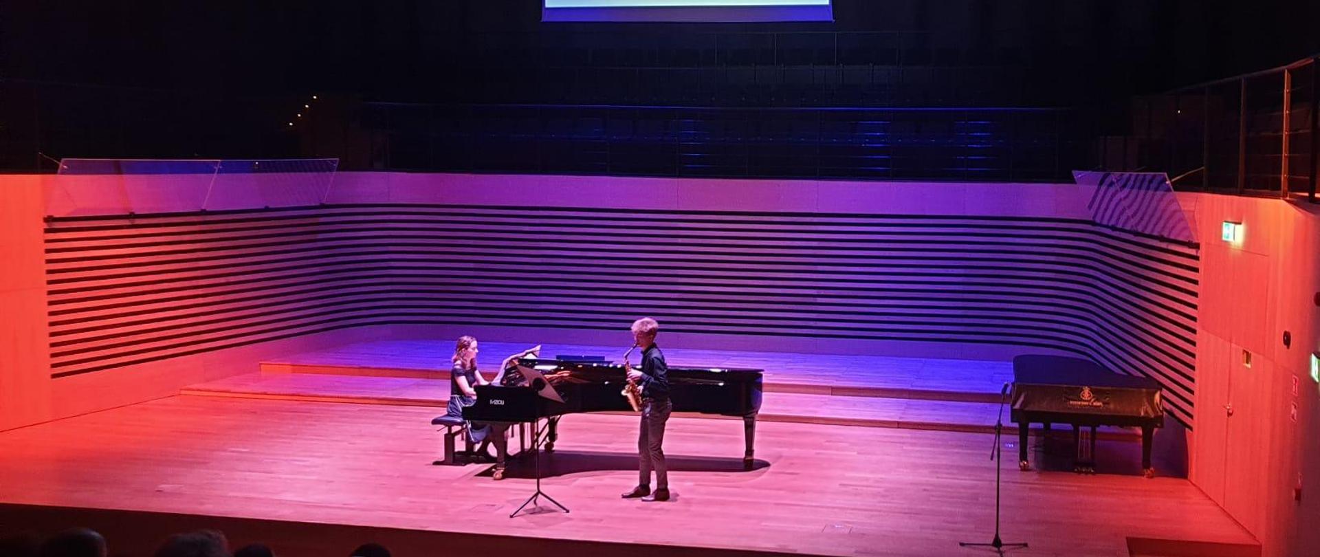 Uczeń gra na saksofonie stojąc przed fortepianem, akompaniatorka gra na fortepianie na scenie sali koncertowej PSM