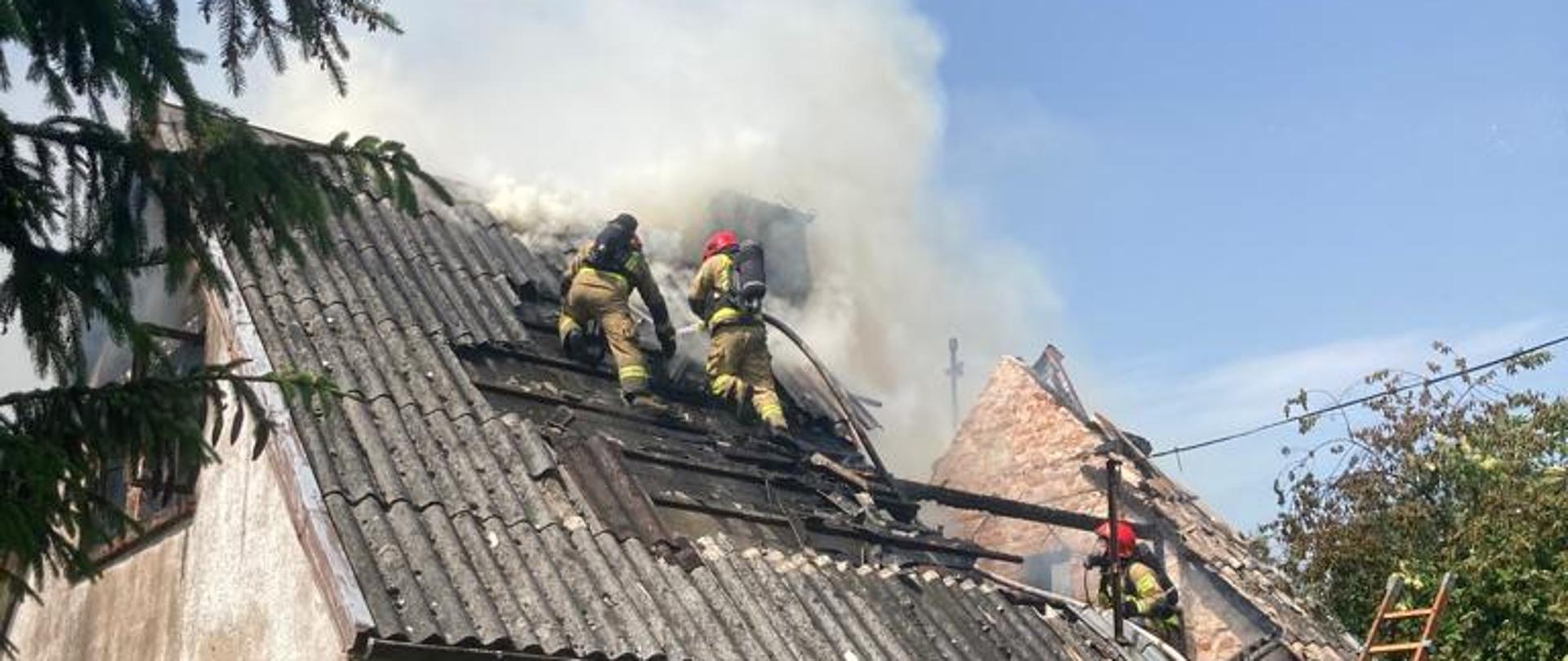 Fotografia przedstawia strażaków gaszących palący się budynek, dwóch znajduje się na dachu, widać dym. Przy budynku widać drabinę strażacką i strażaków na miejscu zdarzenia.