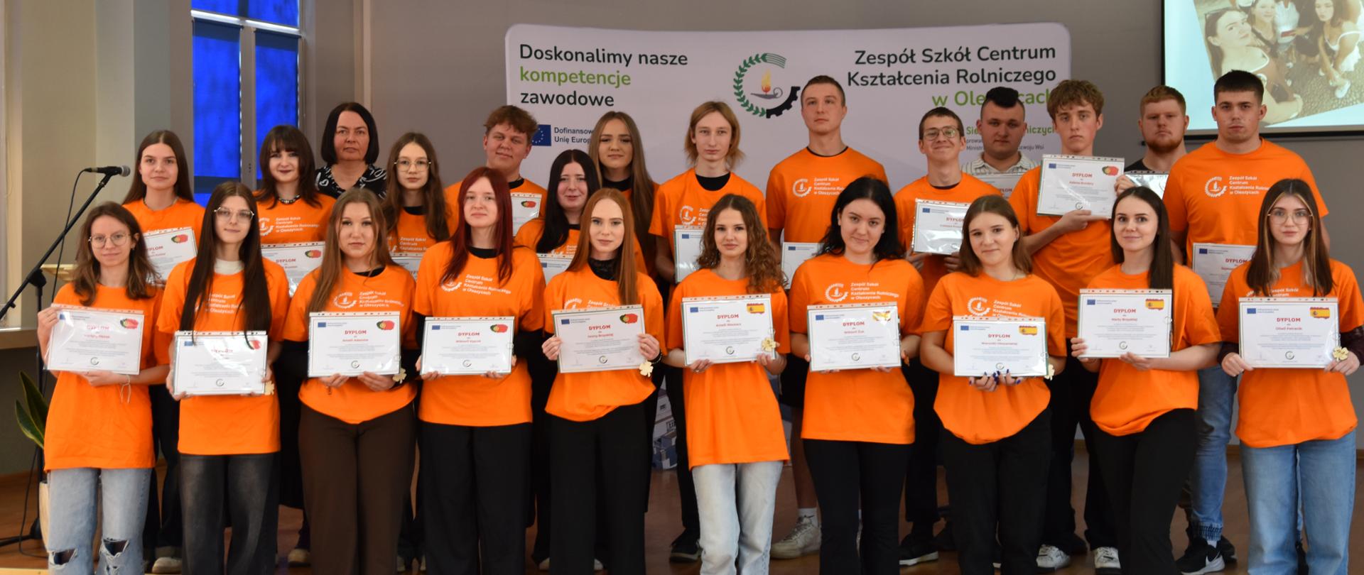 Duże grupa uczniów w pomarańczowych koszulkach stoi przy scenie, każdy trzyma przed sobą certyfikat. Za stojącymi uczniami widać baner z napisem Doskonalimy nasze kompetencje zawodowe logiem i nazwą szkoły ZSCKR w Oleszycach oraz okno zasłonięte granatowa roletą. 