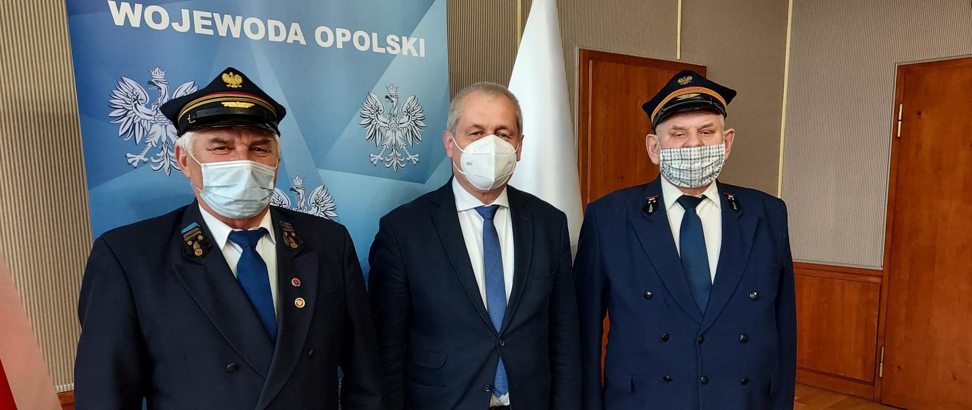 Spotkanie wojewody opolskiego ze Stowarzyszeniem Miłośników Kolei w Racławicach Śląskich. Na zdjęciu trzech mężczyzn, w środku wojewoda opolski a po bokach kolejarze. Mężczyźni ubrani są w ciemne garnitury, na twarzy mają maseczki ochronne. W tle roll up granatowy z napisem Wojewoda Opolski, oraz po lewej i po prawej stronie biało-czerwone flagi. 