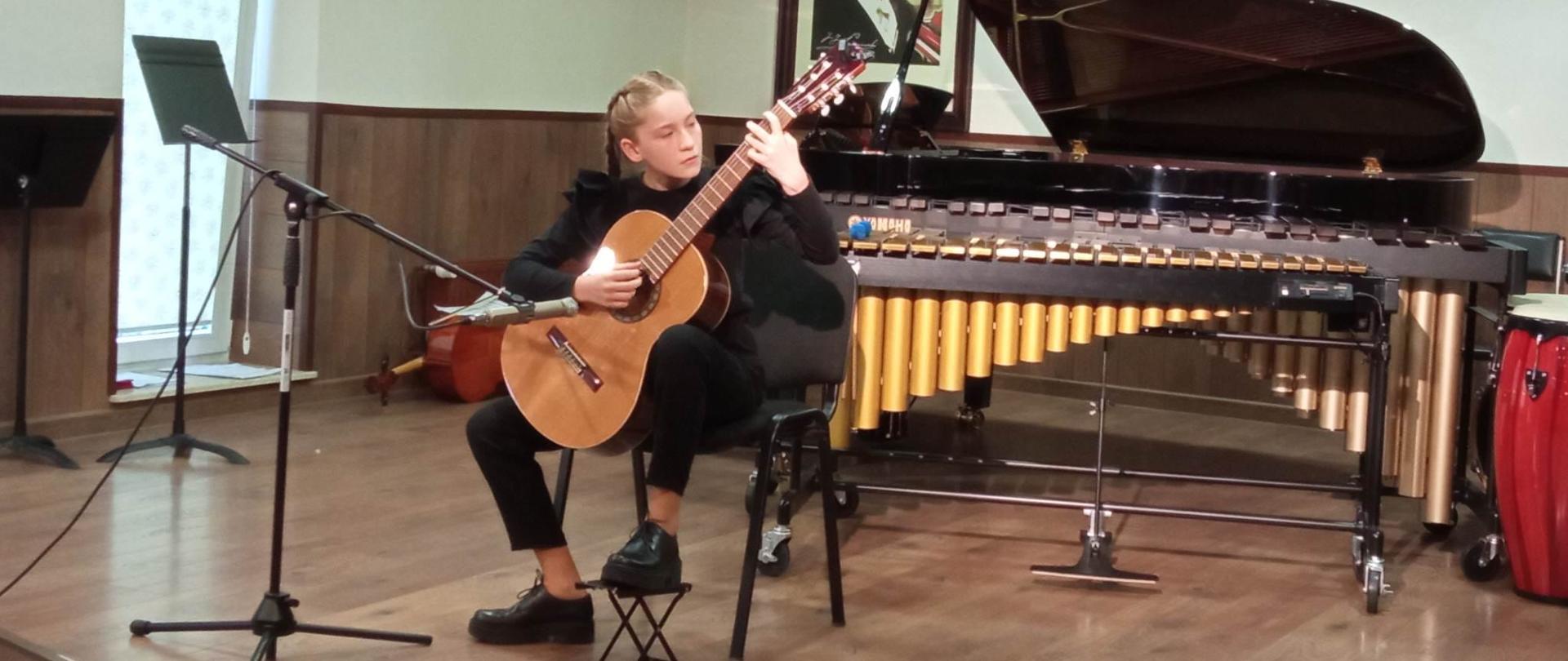 Scena z podłogą koloru brązowego, na środku uczennica ubrana na czarno siedzi ja krześle, gra na gitarze. Przed nią mikrofon na statywie, za nią marimba oraz czarny fortepian. Ściany koloru białego i brązowego. Z lewej strony uchylone okno. z tyłu wisi portret Paderewskiego. Na suficie zapalone okrągłe lampy.