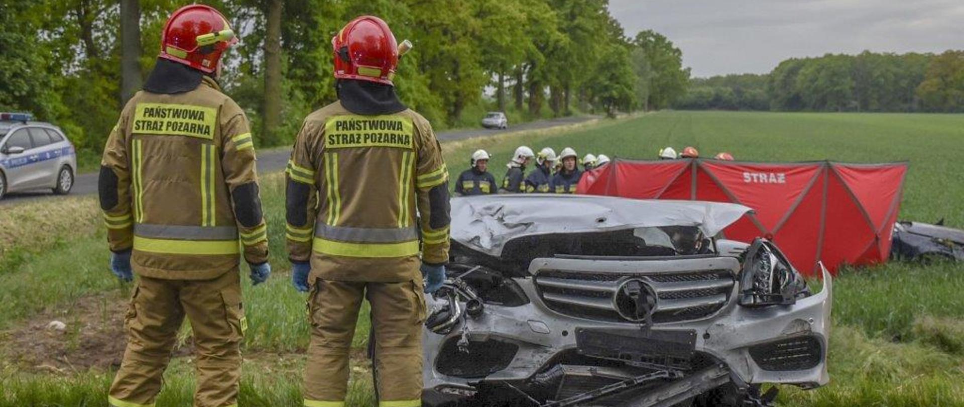 Dwóch strażaków w ubraniach bojowych stoi przed rozbitym pojazdem marki Mercedes. Pojazd ma całkowicie zdeformowany przód. W tle widać parawan ratowniczy, który odizolowuje widok drugiego pojazdu od osób postronnych. Wokół pojazdu znajdują się strażacy z Ochotniczej Straży Pożarnej oraz Państwowej Straży Pożarnej. Drugi z pojazdów znajduje się kilka metrów od drogi na pobliskim polu.