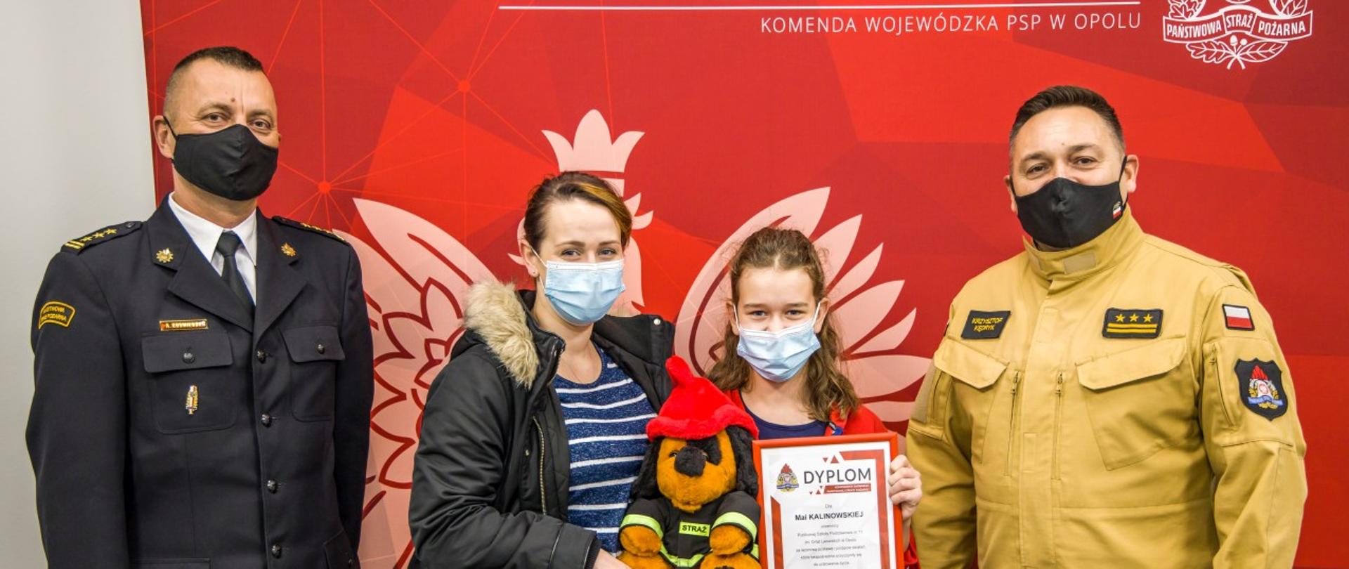 Pamiątkowe zdjęcie Opolskiego Komendanta Wojewódzkiego PSP st. bryg. Krzysztofa Kędryka (umundurowanie służbowe koloru piaskowego) i jego zastępcy st. bryg. Arkadiusza Kuśmierskiego (umundurowanie wyjściowe) z Mają Kalinowską i jej mamą Nicole Walczak. Dziewczynka oraz mam trzymają upominki PSP oraz dyplom Komendanta Głównego PSP. Osoby stoją na tle czerwonej planszy PSP z białym napisem Państwowa Straż Pożarna oraz Komenda Wojewódzka PSP. Na planszy również w jasnym kolorze orzeł.