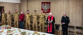 Zdjęcie przedstawia uroczyste spotkanie wielkanocne w Komendzie Miejskiej PSP w Ostrołęce. W sali o ścianach wyłożonych jasnym drewnem, ozdobionej godłem Polski, zgromadzili się strażacy i zaproszeni goście. Większość z nich ma na sobie jasnobrązowe mundury koszarowe z naszywkami z flagą Polski. Pośrodku stoi kapelan w czarnej sutannie i czerwonej stule z wyhaftowanymi krzyżami, czytający z modlitewnika. Po jego lewej stronie stoi Komendant w mundurze wyjściowym z odznaczeniami. Wzdłuż ściany za nimi stoi szereg krzeseł, na których siedzi kilku mężczyzn. Na pierwszym planie widać długi stół przykryty białym obrusem, zastawiony obficie tradycyjnymi potrawami wielkanocnymi. Po lewej stronie stołu widać soki owocowe i ciasto babkę. Atmosfera jest spokojna i uroczysta.