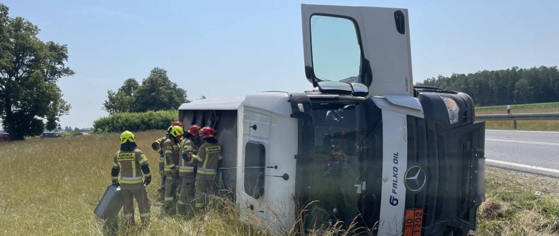 Zdjęcie przedstawia leżący na boku w rowie samochód-cysternę, przewożący olej napędowy. Przy pojeździe znajduje się sześciu strażaków w ubraniu bojowym, zbierających wyciekający z dwóch dekli znajdujących się w górnej części płaszcza zbiornika olej napędowy do kast. Zdjęcie wykonano w słoneczny dzień, przy zjeździe z obwodnicy Zambrowa w kierunku Łomży.