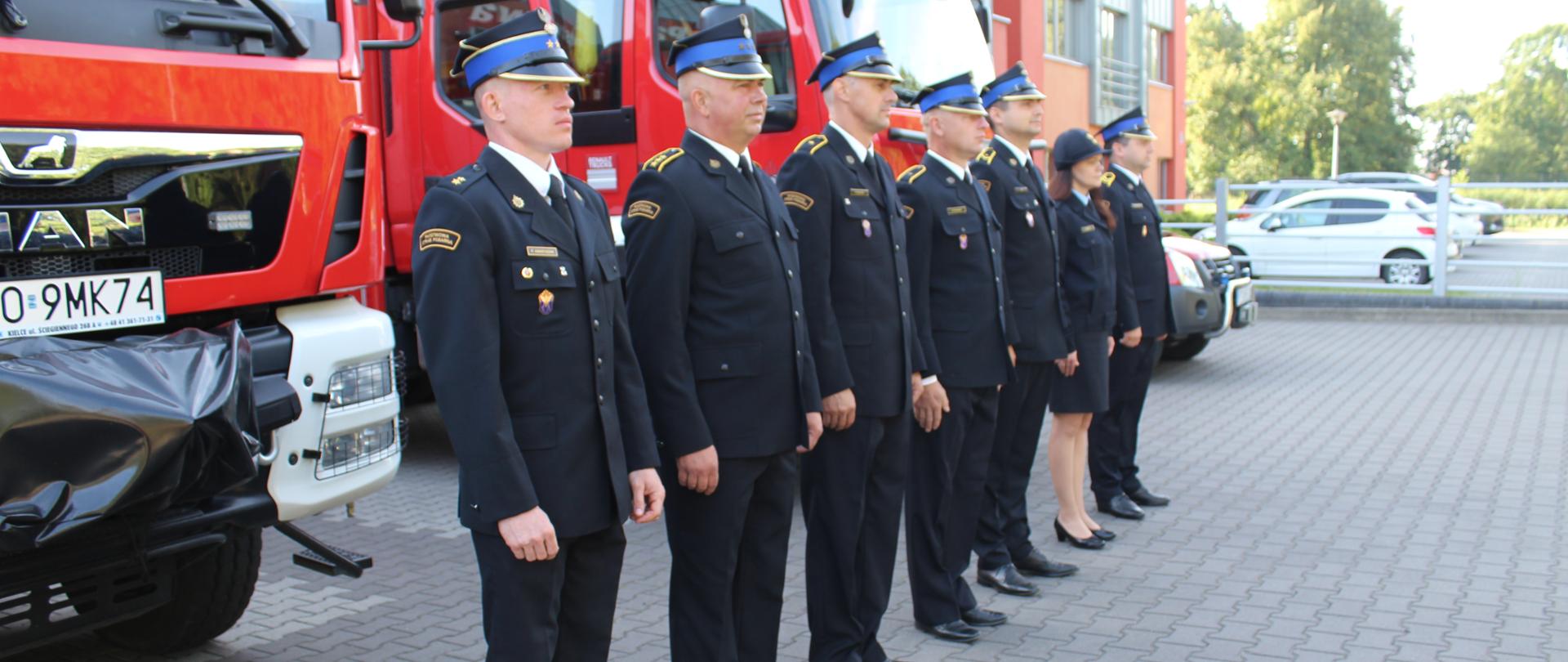 Awansowani na tle samochodów pożarniczych.
