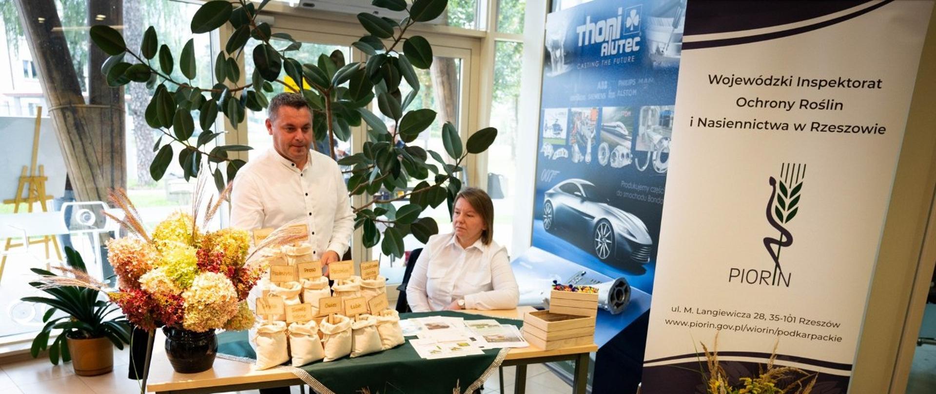 Mężczyzna i kobieta, znajdują się za stołem. Na stole są kwiaty, torebki z nasionami. Przed stołem również poukładane są kolorowe kwiaty, dynie i koszyk z warzywami. Po prawej stronie stoi biało-granatowy baner z logiem instytucji.