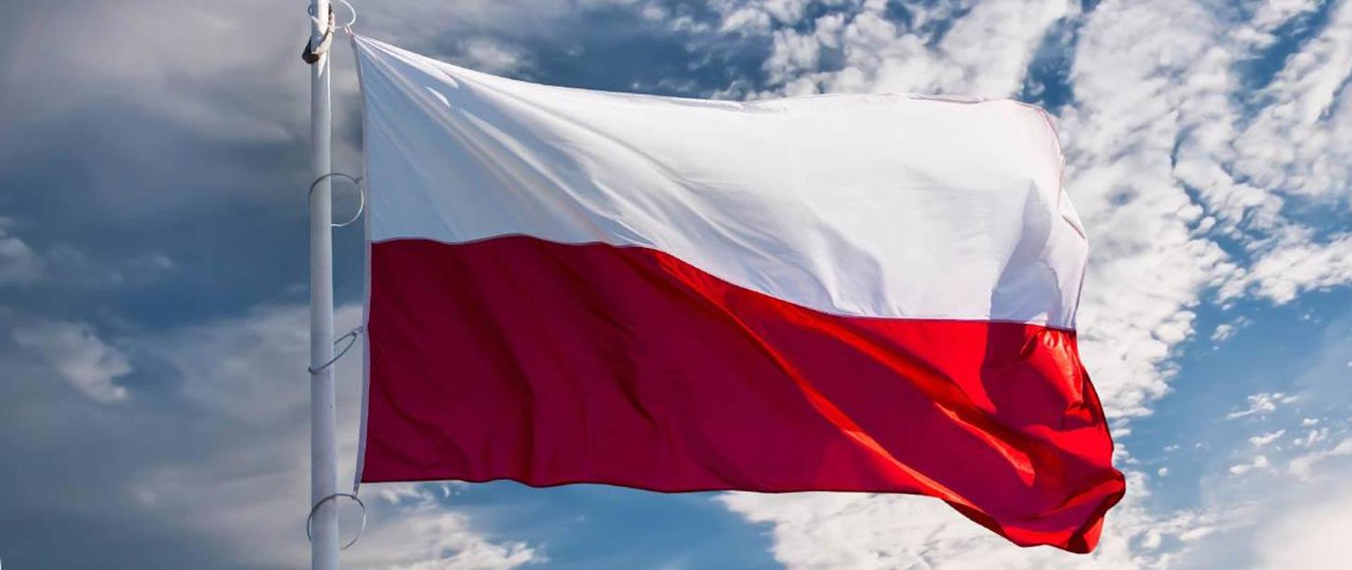 Na pierwszym planie powiewająca flaga Polski zawieszona na maszcie. Tło zdjęcia to słoneczne lekko zachmurzone niebo