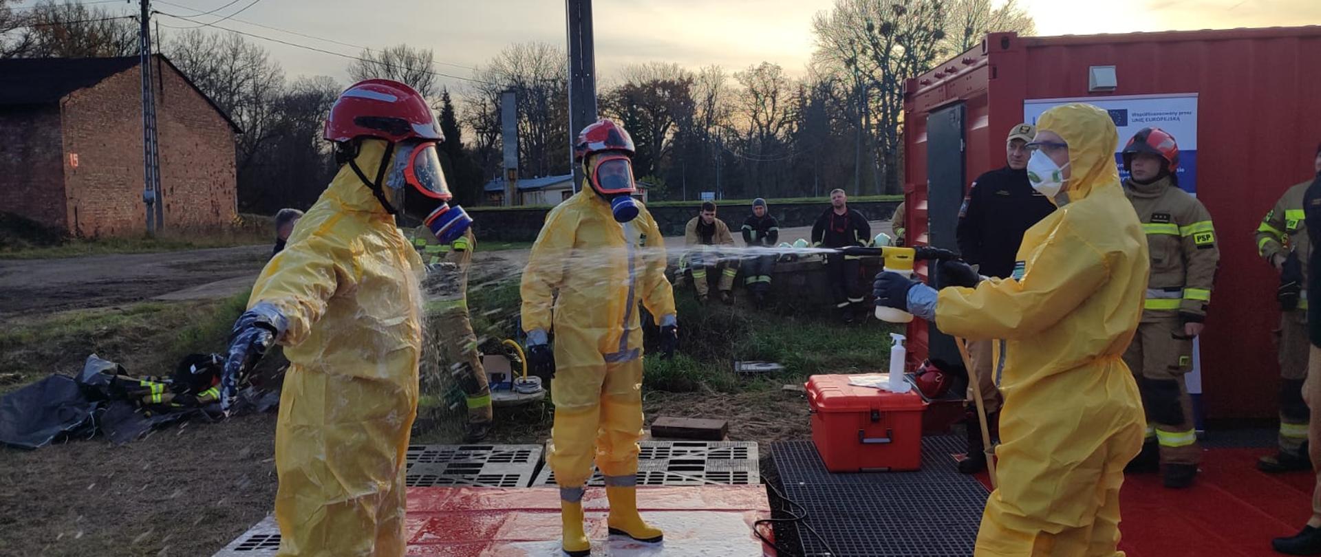 Ratownicy w żółtych kombinezonach ochronnych stoją na czerwonej macie przed czerwonym kontenerem. Jeden z nich pomaga drugiemu zdjąć hełm za pomocą długiego narzędzia. Obok znajduje się tablica informacyjna projektu Interreg oraz skrzynka ze sprzętem.