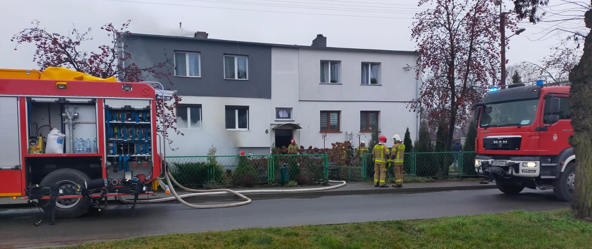 Na zdjęciu widać dwa samochody pożarnicze oraz strażaków. W tle widać dom wielorodzinny w którym powstał pożar