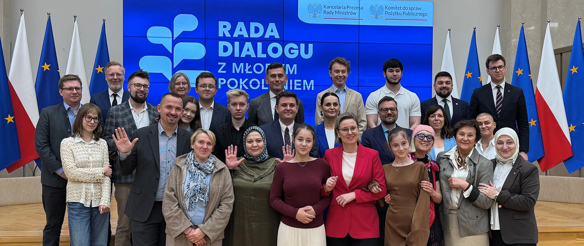 Zdjęcie grupowe. Uczestnicy wydarzenia pozują do wspólnego zdjęcia. W pierwszym rzędzie widoczna między innymi Sekretarz Stanu Adriana Porowska. W tle widoczny ekran, na którym wyświetlone jest logo Rady Dialogu z Młodym Pokoleniem. Po obu stronach ekranu stoją flagi Polski i Unii Europejskiej.