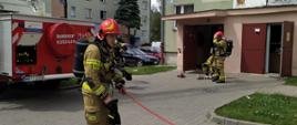 Strażak w ubraniu specjalnym zwija wąż pożarniczy przed budynkiem wielorodzinnym. W tle dwóch strażaków uczestniczących w ćwiczeniach.
