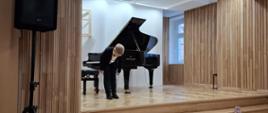 Na zdjęciu widzimy młodego pianistę stojącego na scenie w eleganckim wnętrzu sali koncertowej. Chłopiec, ubrany w ciemną marynarkę i spodnie, pochyla się w geście ukłonu przed publicznością, obok czarnego fortepianu. Podłoga i ściany wyłożone są jasnym, drewnianym parkietem i pionowymi panelami akustycznymi, co podkreśla estetykę i akustykę sali. Wnętrze jest jasne i nowoczesne, a całość oddaje uroczystą i serdeczną atmosferę wydarzenia muzycznego.
