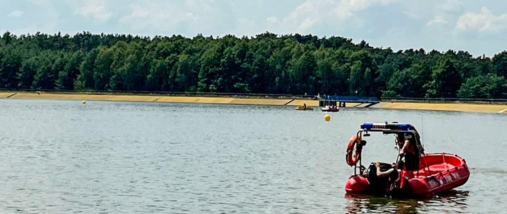 Łódź strażacka ze strażakami pływa po zalewie. W tle las.