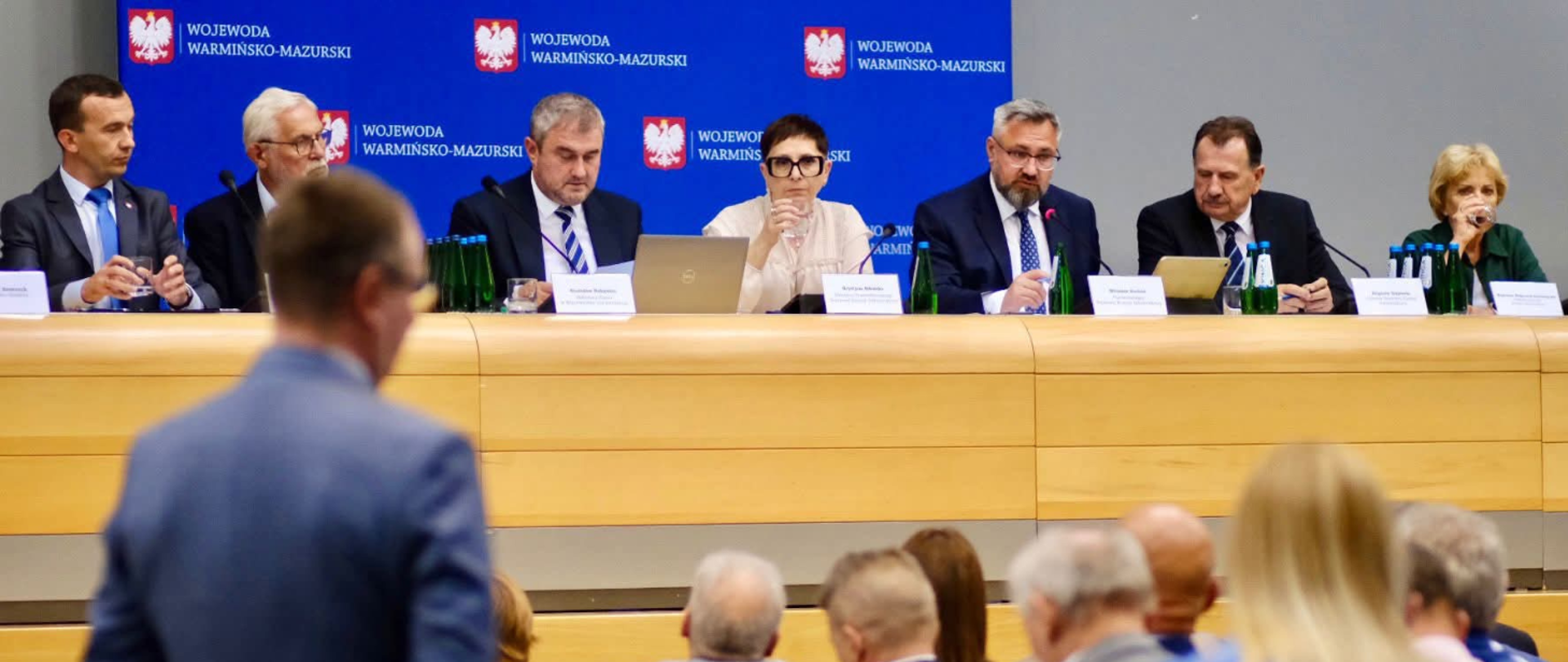 Duży stół, na którym stoją mikrofony oraz komputery, za nim siedzą ludzie. Na przeciwko stołu siedzą ludzie.