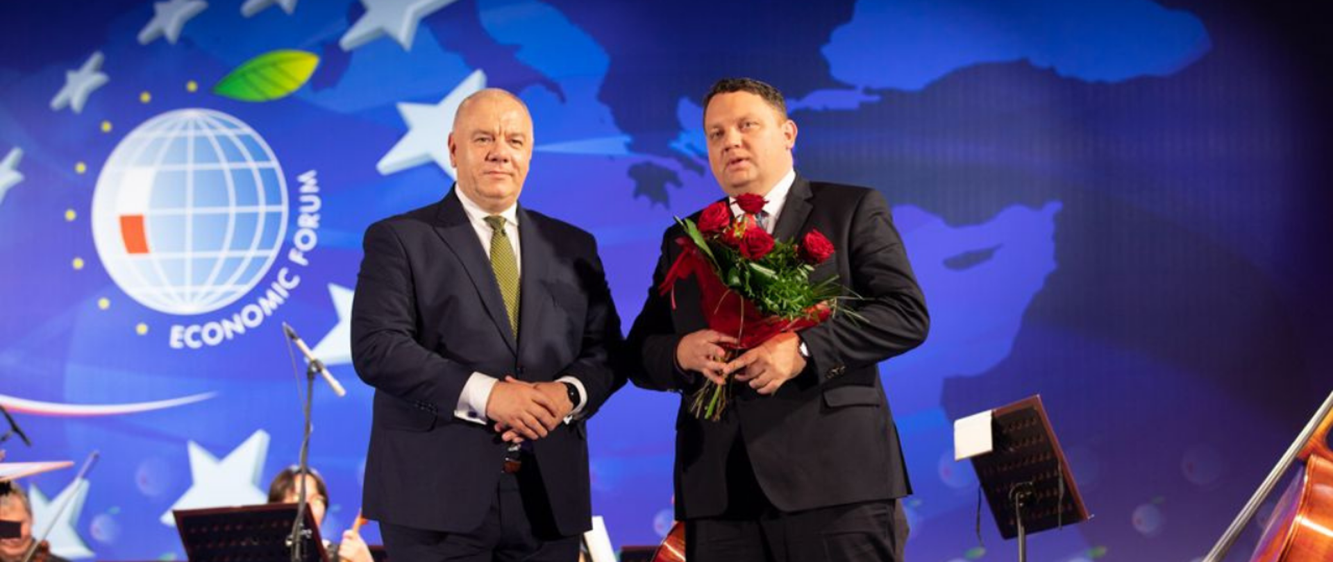 Gala podczas Forum Ekonomicznego. Do zdjęcia pozują wicepremier Jacek Sasin i prezes KGHM Marcin Chludziński. Szef spółki trzyma kwiaty i statuetkę. W tle ścianka z logo imprezy i członkowie orkiestry.