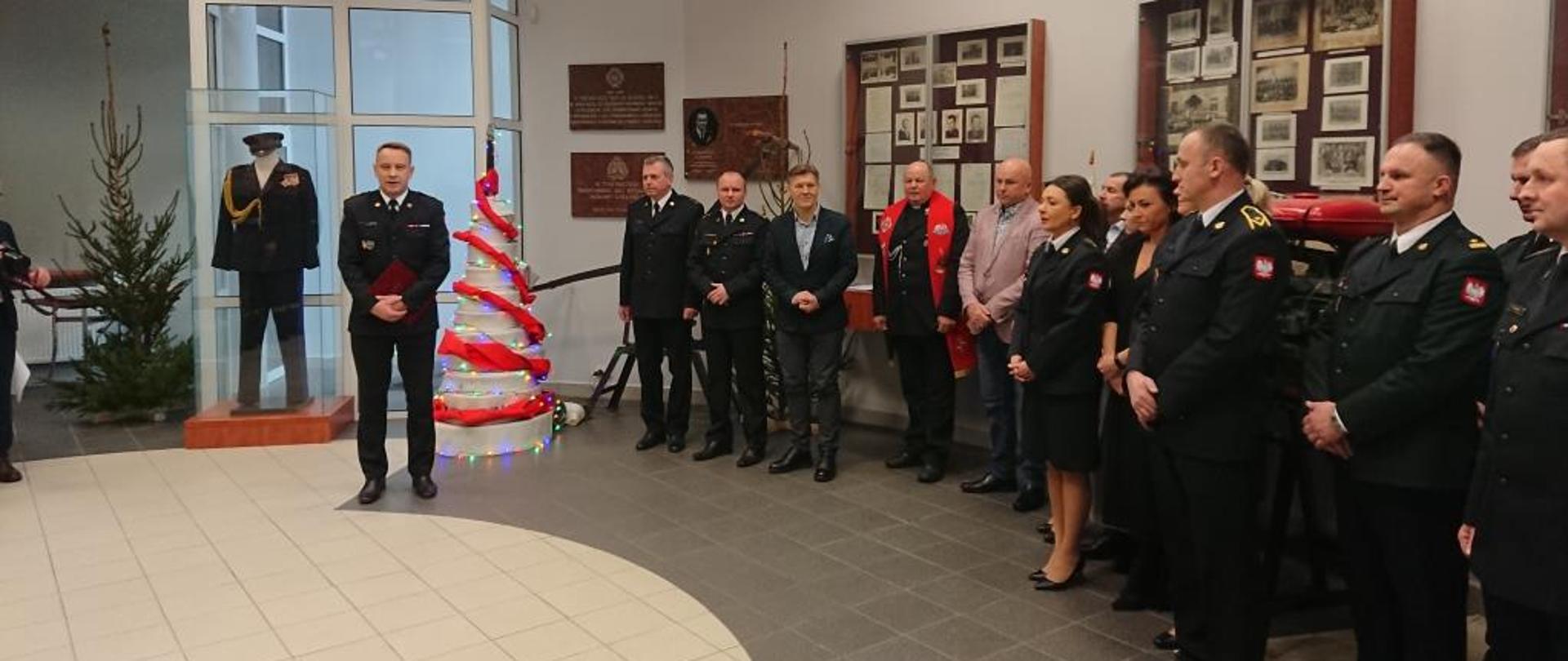 Zdjęcie wykonane w jednym z pomieszczeń komendy miejskiej Państwowej Straży Pożarnej w Radomiu - sali tradycji imienia generała Zygmunta Jarosza. Na zdjęciu widać funkcjonariuszy, księdza kapelana oraz samorządowców. Zebrani uczestniczą w spotkaniu wigilijnym. Przed nimi stoi komendant miejski Państwowej Straży Pożarnej w Radomiu - starszy brygadier Bartosz Musiał. Funkcjonariusz składa zebranym na sali życzenia z okazji zbliżających się Świąt Bożego Narodzenia. 