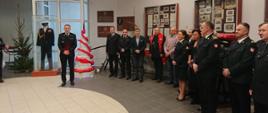 Zdjęcie wykonane w jednym z pomieszczeń komendy miejskiej Państwowej Straży Pożarnej w Radomiu - sali tradycji imienia generała Zygmunta Jarosza. Na zdjęciu widać funkcjonariuszy, księdza kapelana oraz samorządowców. Zebrani uczestniczą w spotkaniu wigilijnym. Przed nimi stoi komendant miejski Państwowej Straży Pożarnej w Radomiu - starszy brygadier Bartosz Musiał. Funkcjonariusz składa zebranym na sali życzenia z okazji zbliżających się Świąt Bożego Narodzenia. 