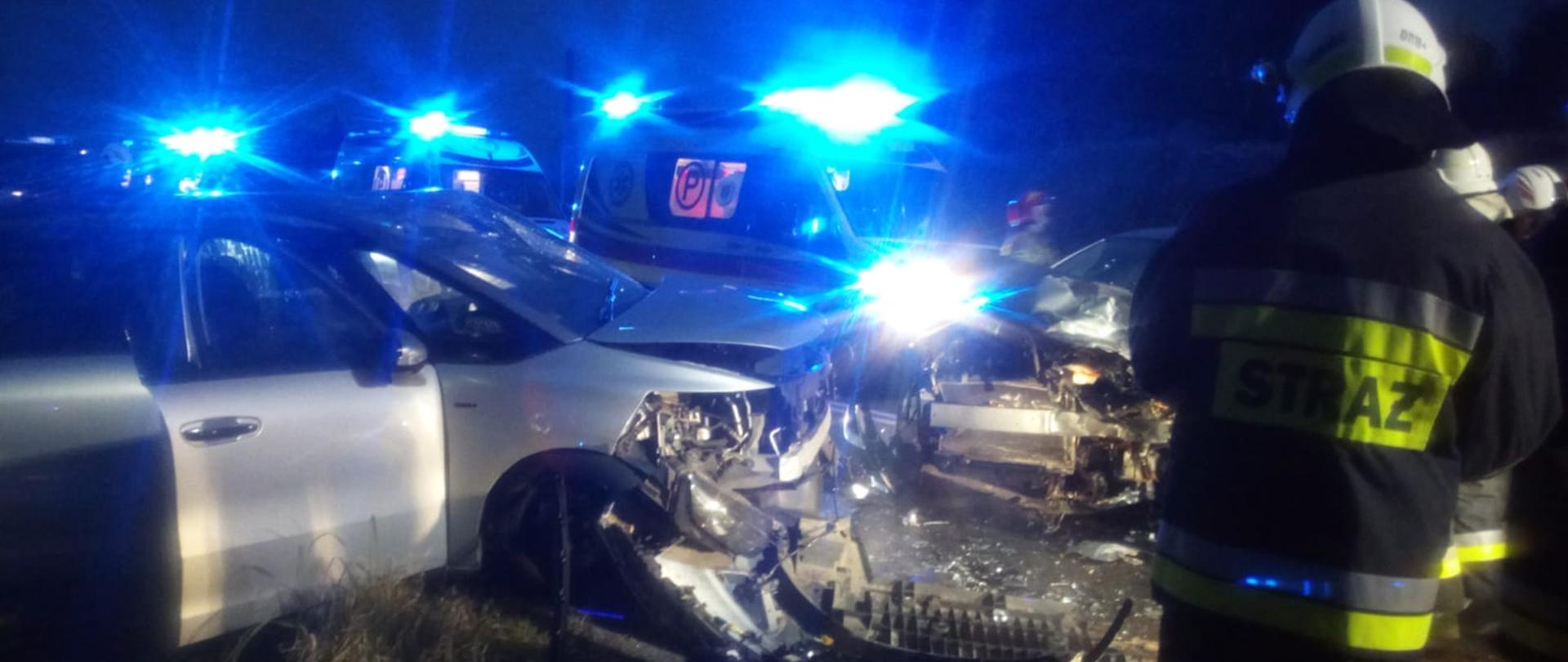 Zdjęcie wykonane w porze nocnej obrazuje zniszczone w wyniku zderzenia samochody osobowe. Jeden z nich znajduje się częściowo na poboczu. Na drodze widoczne porozrzucane rozbite elementy z pojazdów oraz przemieszczający się strażacy. Za zniszczonymi samochodami stoją karetki pogotowia ratunkowego z włączonymi niebieskimi światłami błyskowymi.