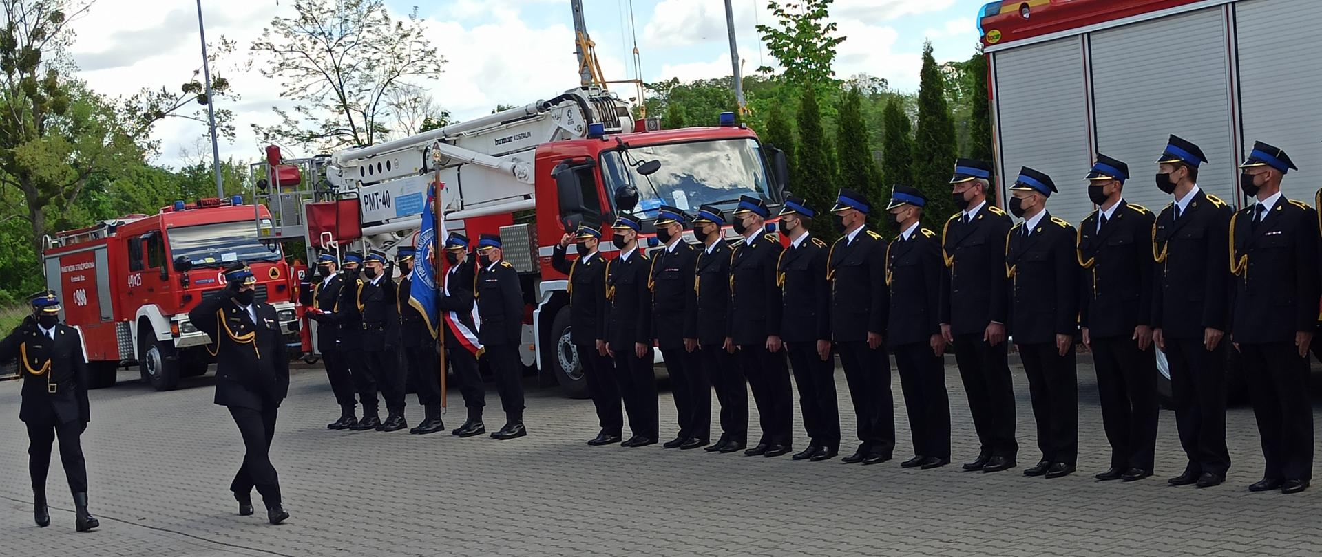 Przed pocztem flagowym, pocztem sztandarowym oraz szeregiem strażaków w mundurach galowych przechodzi przedstawicielem Mazowieckiego Komendanta Wojewódzkiego oraz dowódca uroczystości salutują. W tle widać samochody pożarnicze.