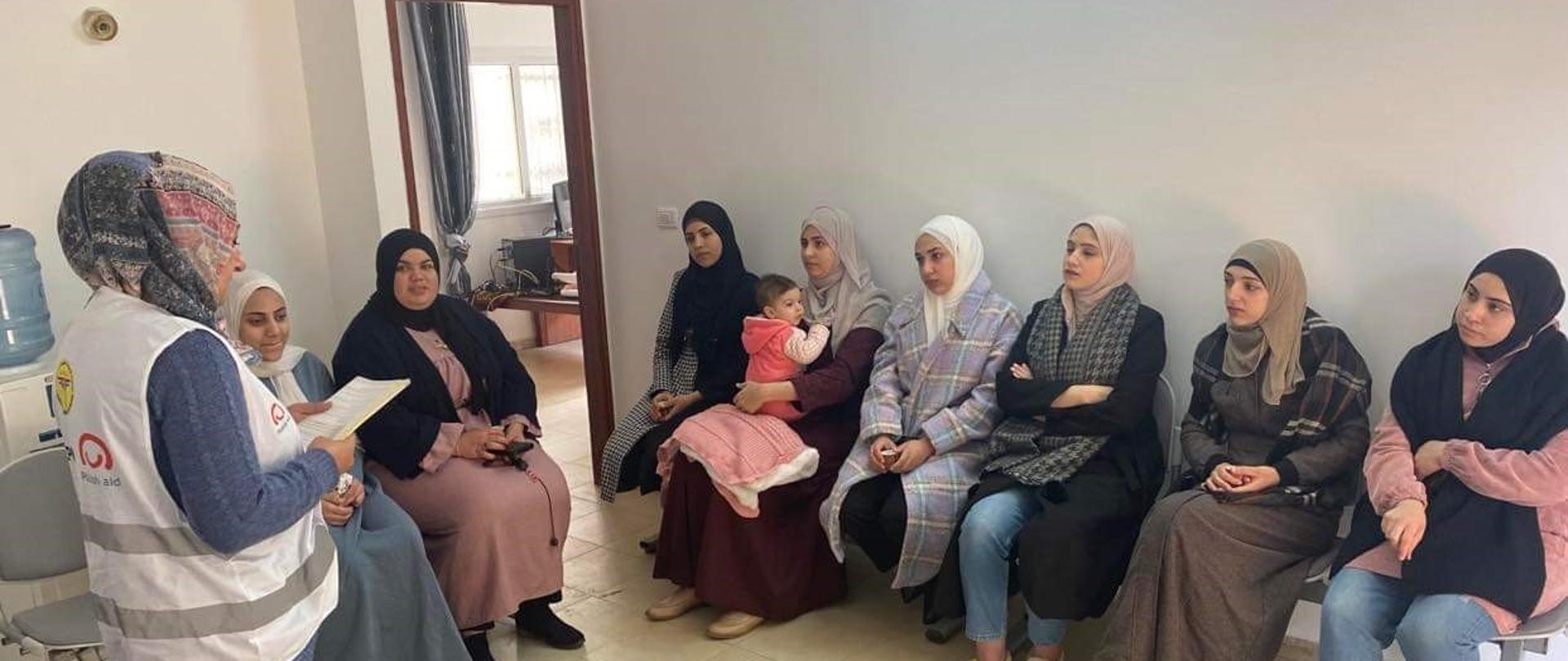 Kobiety z głowami ubranymi w chusty siedzą na krzesłach pod ścianą. Przed nimi stoi kobieta, również z głową zasłoniętą chustą i patrzy się na kartki papieru, które trzyma przed sobą. 