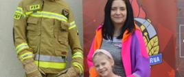 Dzień otwarty w rawickiej komendzie. Mama z córką pozują do pamiątkowego zdjęcia na tle logo PSP i manekina-strażaka. Kobieta ma kolorowy płaszczyk, a na głowie przeciwsłoneczne okulary. 