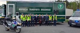 Inspektorzy małopolskiej Inspekcji Transportu Drogowego wraz z przedstawicielami Europejskiego Urzędu ds. Pracy (ELA) stoją na tle Mobilnej Jednostki Diagnostycznej. Obok stoi również oznakowany motocykl i oznakowany furgon małopolskiej Inspekcji Transportu Drogowego.