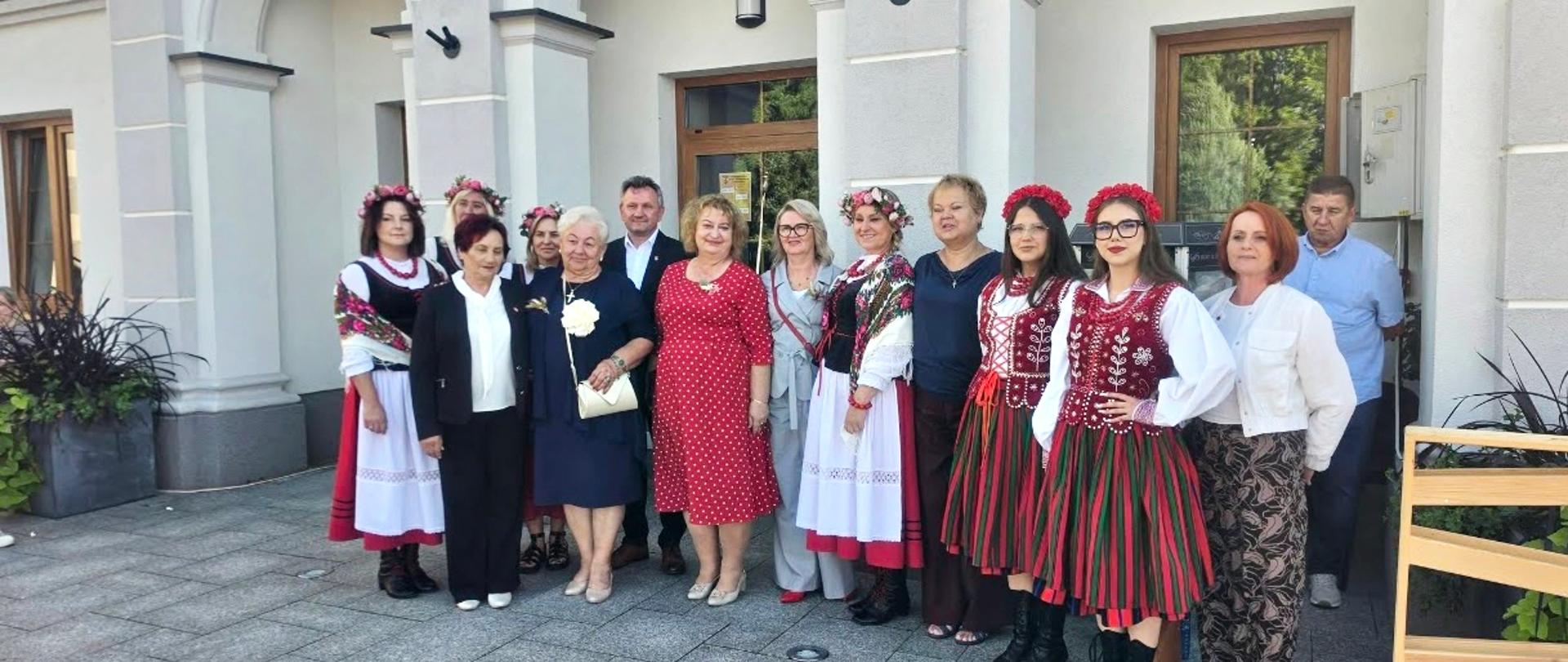 Pamiątkowe zdjęcie grupowe uczestników uroczystości pożegnania wakacji przed budynkiem Centrum Kultury. Na zdjęciu dyrektor placówki, zaproszeni goście oraz uczennice w tradycyjnych strojach ludowych i wiankach.