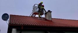 Dwóch strażaków w ubraniach ochronnych pracuje na dachu domu jednorodzinnego z czerwonej dachówki, przy kominie. Jeden stoi na drabinie, drugi w koszu podnośnika. Pochmurne niebo w tle.