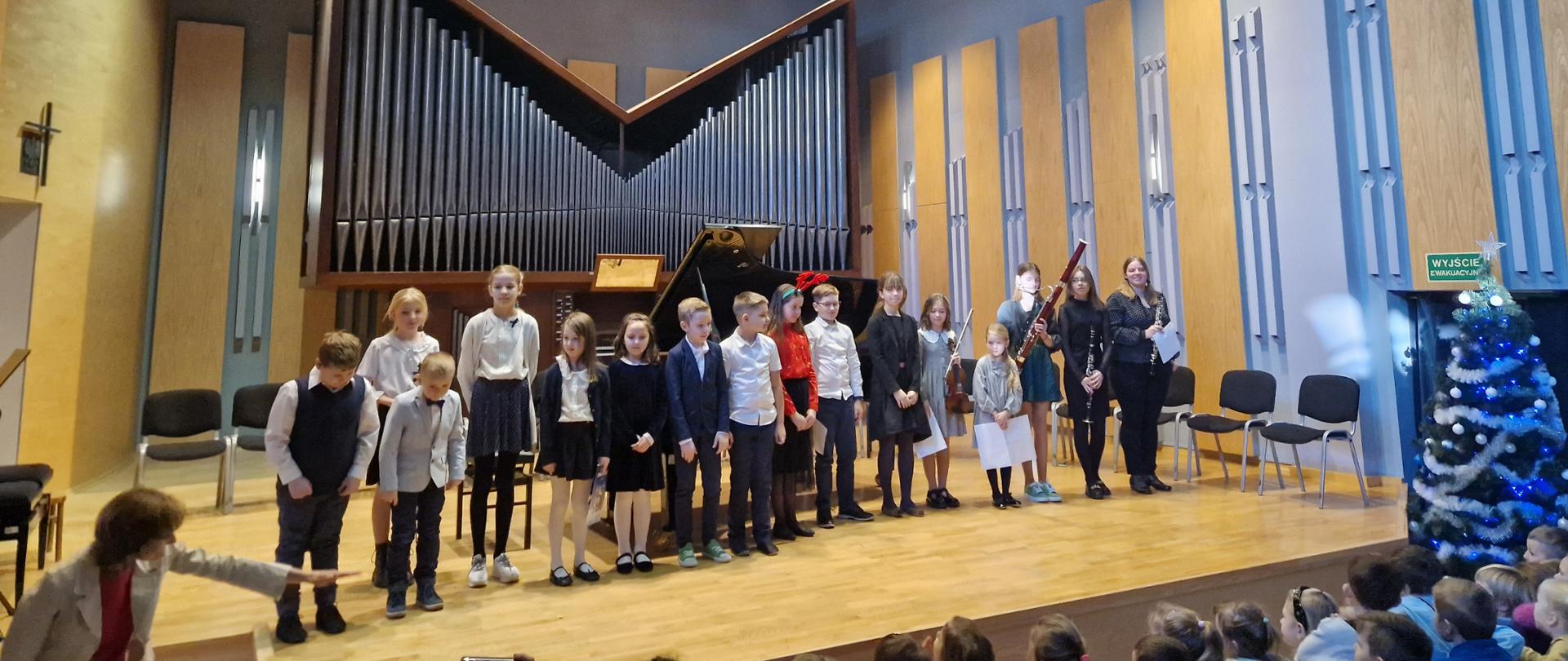 Zdjęcie w sali koncertowej. Na pierwszym planie publiczność złożona z małych dzieci. na scenie sali koncertowej w jednym rzędzie wszyscy uczestnicy koncertu edukacyjnego. Część dzieci trzyma w swoich dłoniach swoje instrumenty. Z lewej strony nauczycielka prowadząca koncert wskazuje lewą ręką na uczestników wydarzenia. Na scenie sali koncertowej krzesła ułożone w półokręgu, Za dziećmi fortepian. a dalej w tle organy, Z lewej strony przed sceną choinka bożonarodzeniowa udekorowana w łańcuchy i bombki.