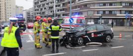 Na zdjęciu widoczny jest pojazd biorący udział w zdarzeniu, ma rozbity przód, obok pracują strażacy, w tle pojazdy straży, pogotowia, policji i budynek biurowy