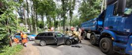 Na zdjęciu strażacy pracują przy uszkodzonym samochodzie osobowym, który zderzył się z pojazdem ciężarowym. Maska auta jest otwarta, a obok leży narzędzie hydrauliczne. W tle widoczne są służby ratownicze, osoby poszkodowane oraz zaparkowane wozy strażackie.