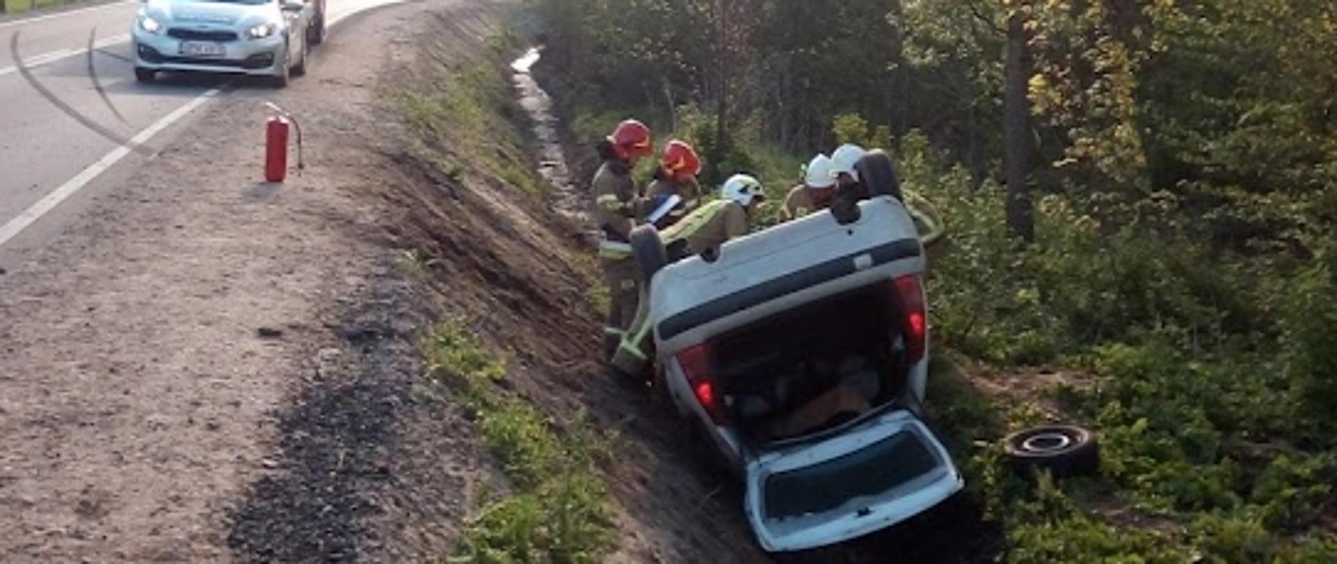 Samochód osobowy leżący na dachu w przydrożnym rowie. Przy samochodzie pracujący strażacy. W tle radiowóz oraz samochód ratowniczo-gaśniczy.