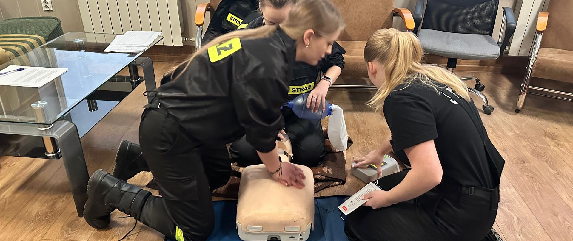 Trzy strażaczki ćwiczą RKO na manekinie w pozycji klęczącej. Jedna uciska klatkę piersiową, a druga używa defibrylatora.