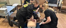 Trzy strażaczki ćwiczą RKO na manekinie w pozycji klęczącej. Jedna uciska klatkę piersiową, a druga używa defibrylatora.