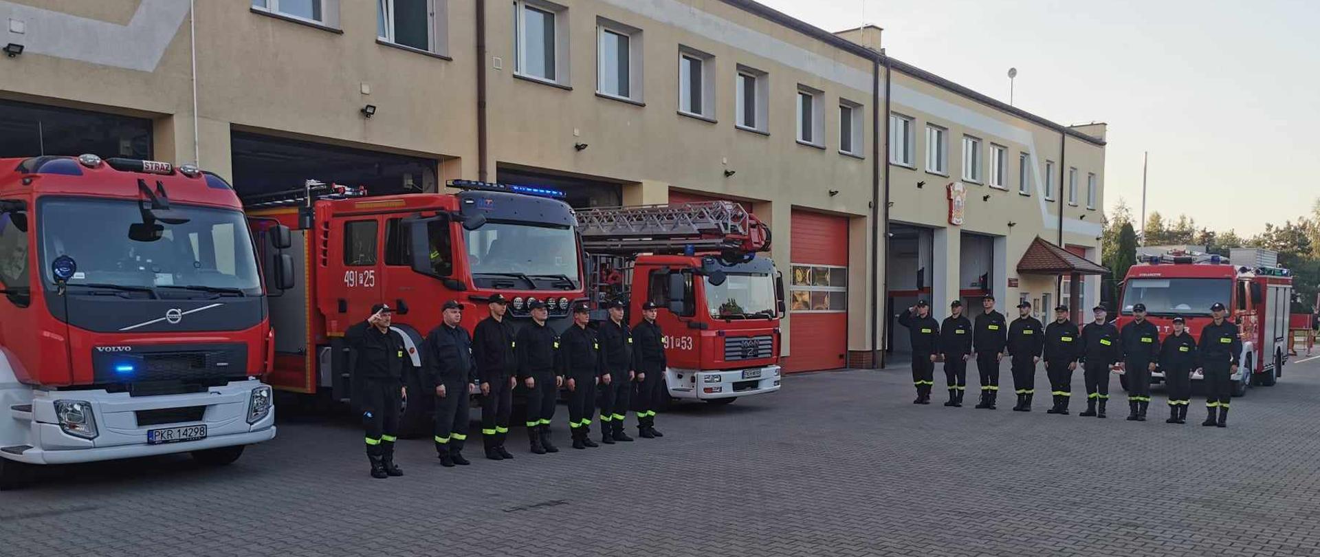 Zdjęcie przedstawia stojących strażaków w umundurowaniu koszarowym na tle samochodów pożarniczych koloru czerwonego. Strażacy oddają hołd zmarłemu tragicznie druhowi.