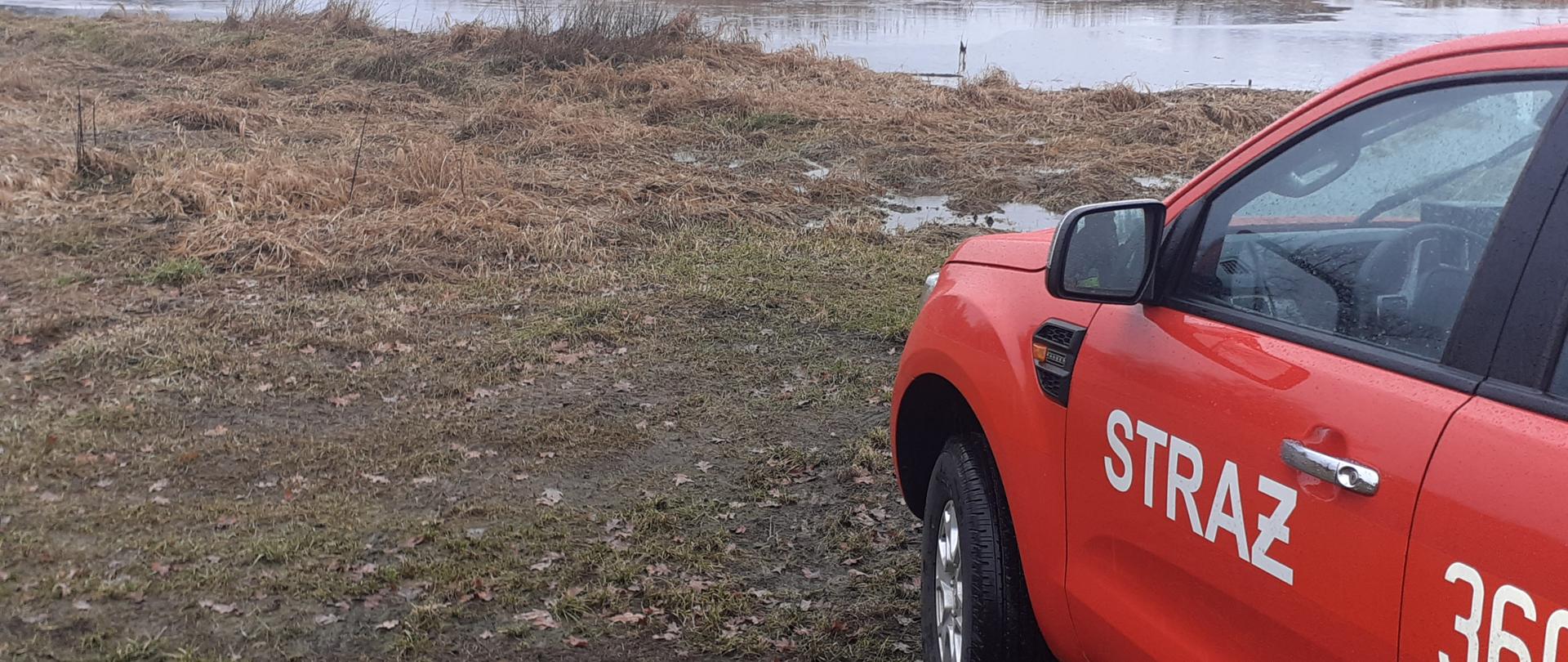 Zdjęcie przedstawia zalane grunty rolne znajdujące się w miejscowości Radnica. Na pierwszym planie znajduje się czerwony samochód osobowy z napisem Straż na drzwiach.