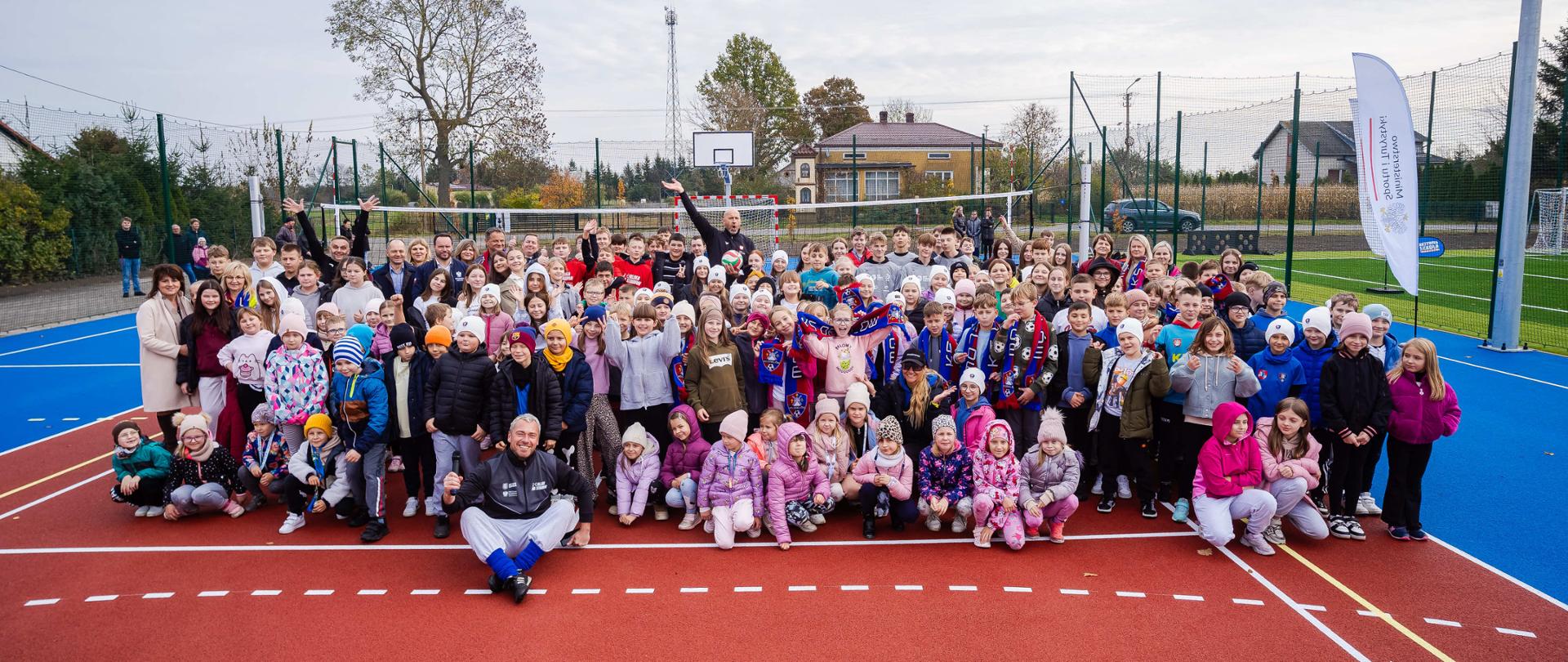 Zdjęcie zbiorowe na Orliku w Drelowie. Na zdjęciu widać dużą grupę dzieci, w ostatnim rzędzie na środku minister sportu i turystyki Jakub Rutnicki, z lewej strony z tyłu wójt gminy Drelów Piotr Kazimierski i wojewoda lubelski Krzysztof Komorski. Z przodu na ziemi siedzi ambasador Aktywnej Szkoły Grzegorz "Gabor" Jędrzejewski. Za fotografowaną grupą widać siatkę do siatkówki, a jeszcze dalej bramkę piłkarską i kosz to koszykówki. Z prawej strony dwa windery z logotypami Ministerstwa Sportu i Turystyki. W tle widać domy i drzewa.