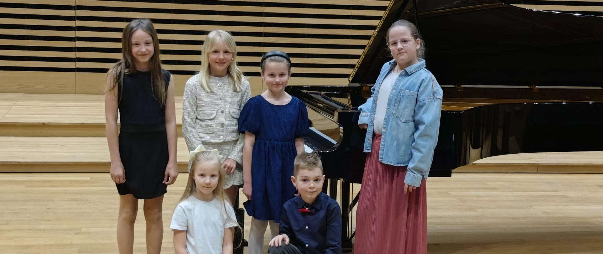 Pięć dziewczynek i chłopiec stoją na scenie przy fortepianie.