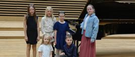 Pięć dziewczynek i chłopiec stoją na scenie przy fortepianie.
