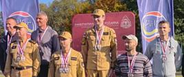 Grupa uczestników wydarzenia sportowego pozuje do wspólnego zdjęcia przy podium z oznaczeniami drugiego i trzeciego miejsca. Część osób ubrana jest w umundurowanie, a uczestnicy mają na szyjach medale. Przed podium znajduje się sprzęt sportowy, a w tle widoczne są banery i teren obiektu sportowego.