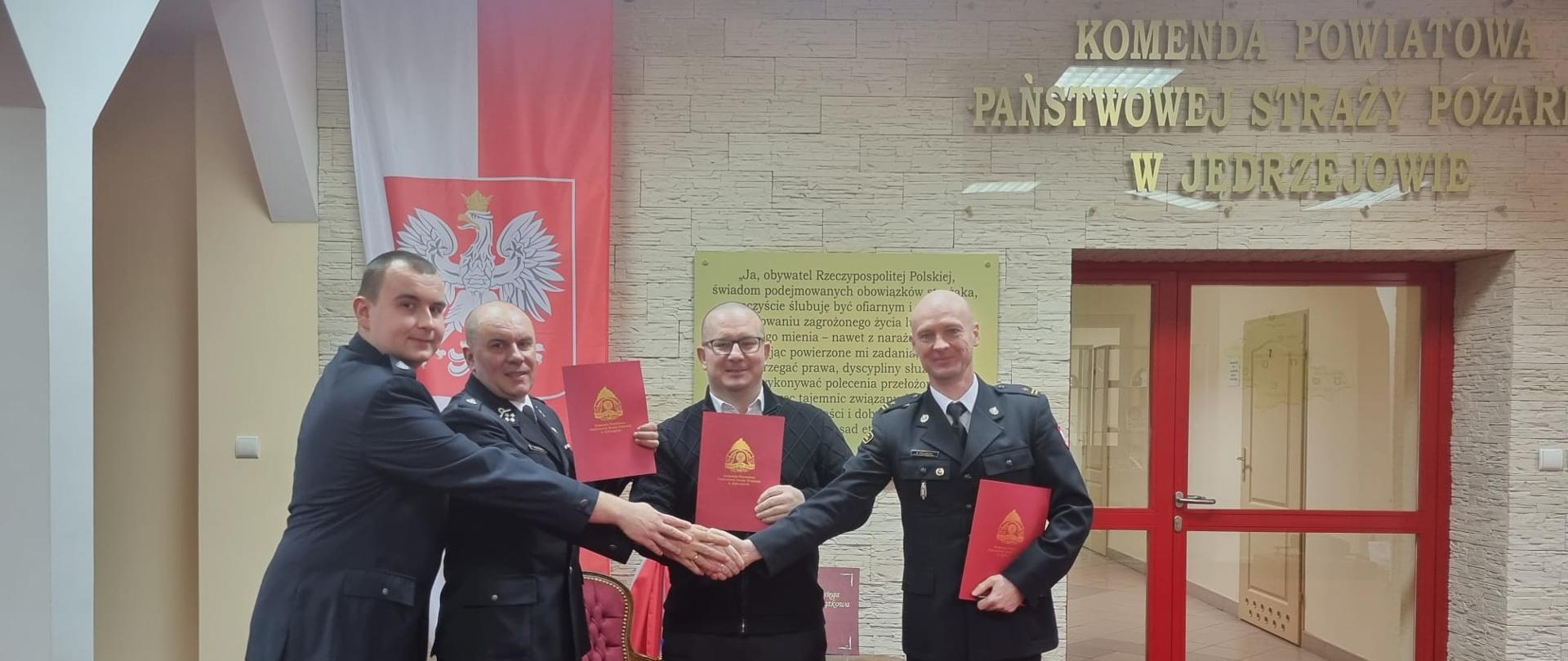 Na zdjęciu widnieje czterech mężczyzn znajdujących się na Sali Tradycji Komendy Powiatowej PSP w Jędrzejowie. Mężczyźni trzymają w rękach podpisane porozumienie dotyczące włączenie jednostki Ochotniczej Straży Pożarnej w Zagaju w struktury krajowego systemu ratowniczo-gaśniczego oraz podają sobie dłonie.