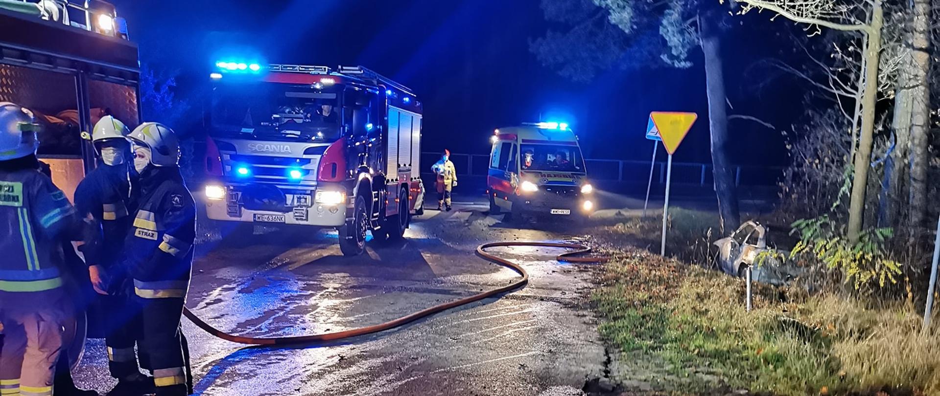 spalony samochód osobowy znajdujący się w rowie - pora nocna, z lewej strony pojazdy służb ratowniczych straży oraz pogotowia ratunkowego